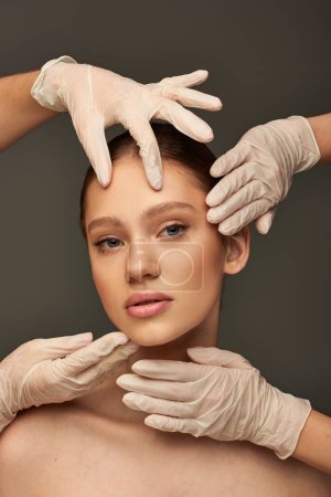 estheticians in medical gloves examining face of young patient on grey background, facial treatment
