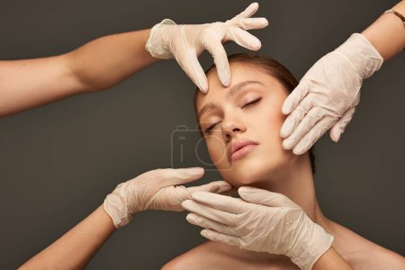 estheticians in medical gloves examining face of young woman with closed eyes on grey background