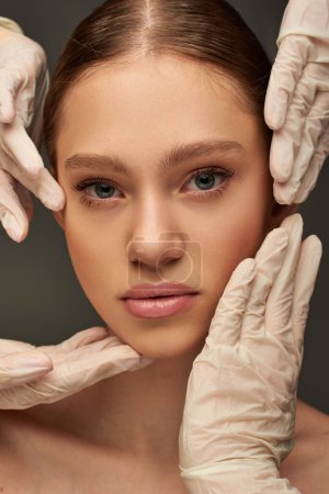 close up of estheticians in medical gloves examining face of young pretty patient on grey background