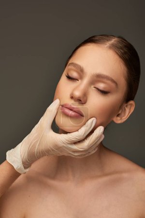Ästhetikerin im Medizinhandschuh berührt Gesicht einer hübschen Frau auf grauem Hintergrund, dermatologisches Konzept
