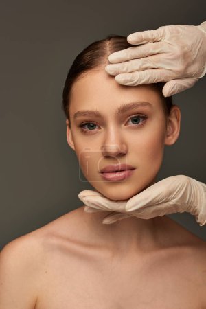 esteticista en guantes médicos tocando la cara de mujer joven sobre fondo gris, concepto dermatológico