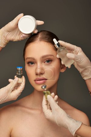estheticians in medical gloves holding different beauty treatment products near young woman on grey