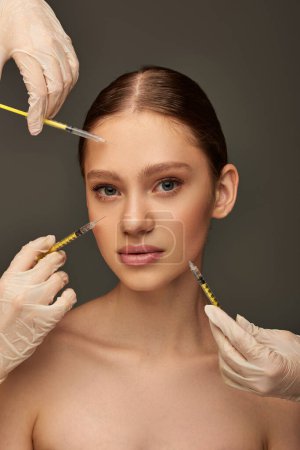 estheticians in medical gloves holding syringes near young woman on grey background, collagen