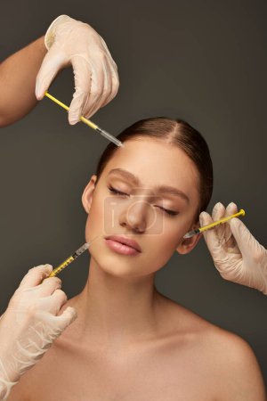 estheticians in medical gloves holding syringes near young woman on grey background, rejuvenation