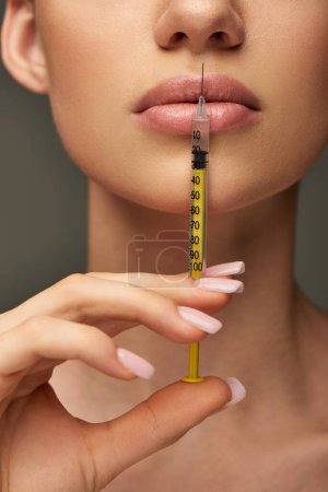 cropped view of young woman holding syringe near face on grey background, lip enhancement concept