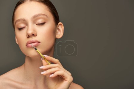 Foto de Retrato de una mujer joven y bonita sosteniendo la jeringa cerca de la cara sobre fondo gris, concepto de realce de labios - Imagen libre de derechos