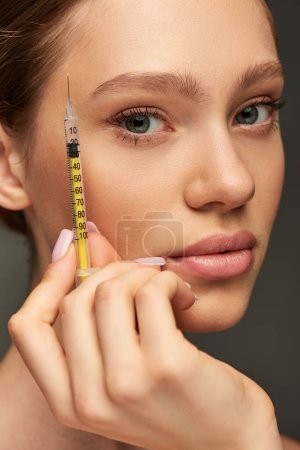 portrait de jeune femme tenant une seringue près du visage sur fond gris, concept de rajeunissement de la peau