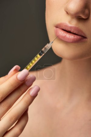 abgeschnittene Aufnahme einer jungen Frau mit Spritze in Gesichtsnähe auf grauem Hintergrund, Konzept zur Lippenvergrößerung