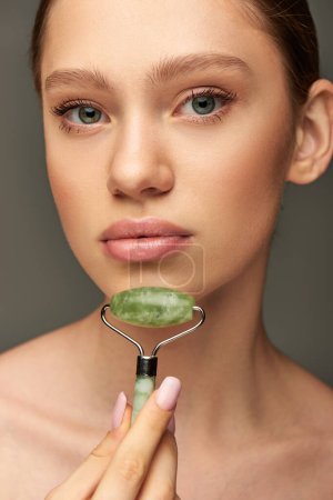 young woman doing face massage with green jade roller on grey background, skin rejuvenation