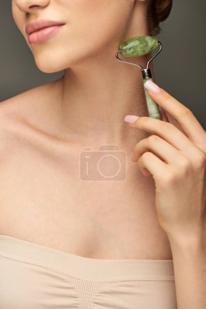 cropped view of woman doing neck massage with jade roller on grey background, rejuvenation