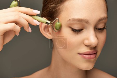 Porträt einer jungen Frau bei einer Gesichtsmassage mit grüner Jadewalze auf grauem Hintergrund, Selbstpflege