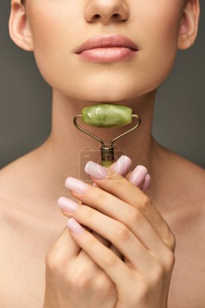 cropped young woman doing massage of neck with green jade roller on grey background, self-care