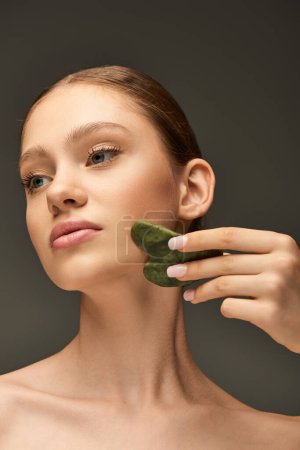 mujer joven haciendo masaje facial con piedra de jade gua sha sobre fondo gris, concepto de contorno facial