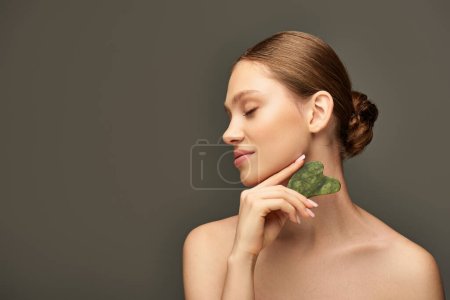 mujer joven haciendo masaje facial con piedra de jade gua sha sobre fondo gris, concepto de cuidado facial