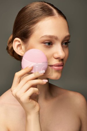 mujer joven que sostiene el dispositivo de cuidado de la piel cerca de la cara sobre fondo gris, cepillo de limpieza rosa