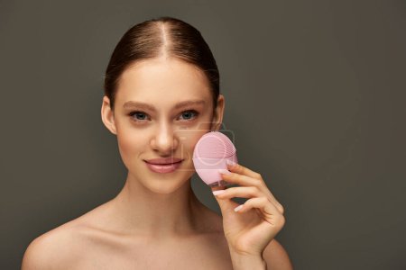 junge Frau mit tragbarem Reinigungspinsel auf grauem Hintergrund, Beauty-Gadget und Hautpflegekonzept