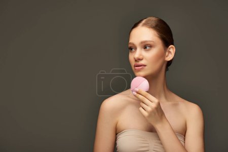 mujer atractiva sosteniendo cepillo de limpieza sobre fondo gris, gadget de belleza y el concepto de cuidado de la piel