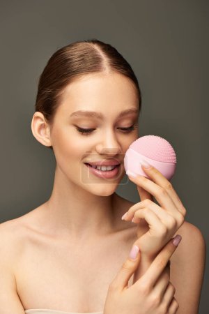 fröhliche Frau mit Reinigungsbürste auf grauem Hintergrund, Beauty-Gadget und Hautpflegekonzept
