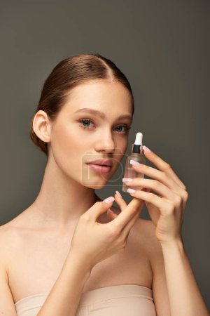 mujer joven con botella de piel brillante con suero sobre fondo gris, hidratación y cuidado de la piel