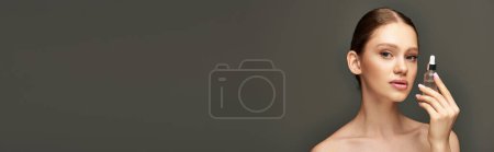 young woman with glowy skin holding bottle with serum on grey background, skin care banner