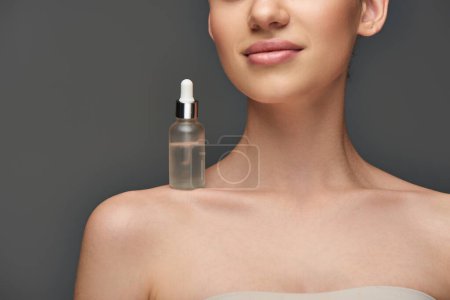 cropped young woman with glowy skin and holding bottle with serum on shoulder on grey background