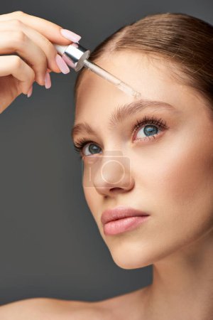 young woman with glowy skin holding dropper with serum on grey background, hydration and skin care puzzle 683988146