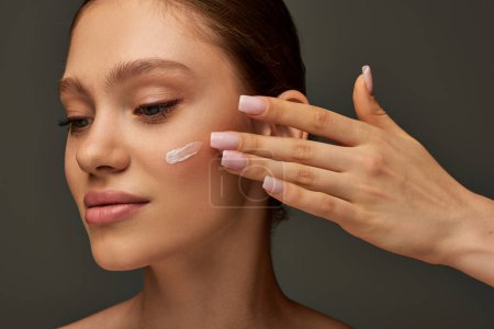 beautiful young woman with bare shoulders applying cream on face on grey background, look away