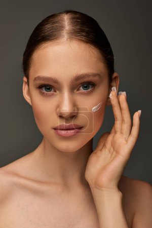 beautiful young woman with bare shoulders applying cream on face on grey background, look at camera