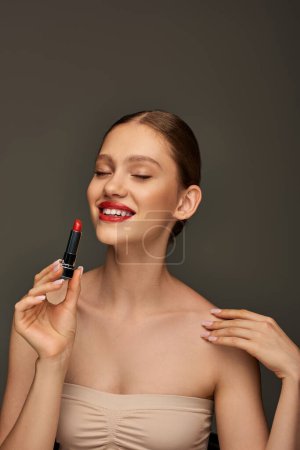 fröhliche junge Frau mit roten Lippen, Lippenstift in der Hand und Lächeln auf grauem Hintergrund, Feiertags-Make-up