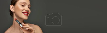 cheerful young woman with red lips holding lipstick and smiling on grey background, makeup banner