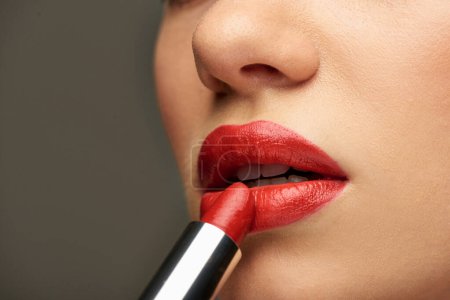 primer plano de mujer joven glamorosa aplicando lápiz labial rojo sobre fondo gris, belleza y maquillaje