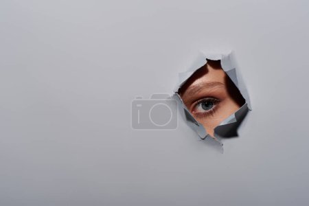 recortado disparo de mujer joven con ojo azul mirando a la cámara a través del agujero en el fondo gris rasgado