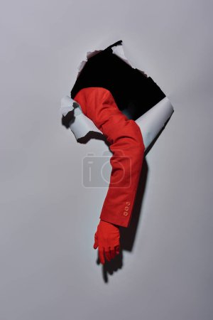 cropped view of woman with red sleeve and glove breaking though hole in grey background, conceptual
