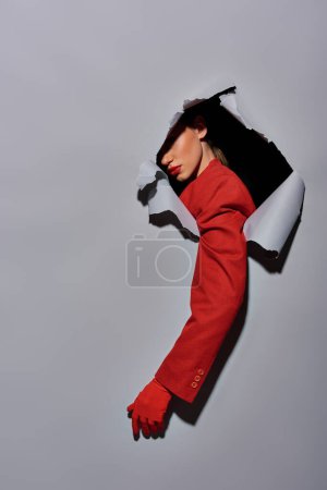Schnappschuss einer Frau mit rotem Ärmel und Handschuh, die durch ein Loch im grauen Hintergrund bricht, konzeptionell