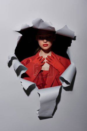 Foto de Atractiva joven con maquillaje audaz en guantes rojos posando cerca del agujero en fondo gris - Imagen libre de derechos