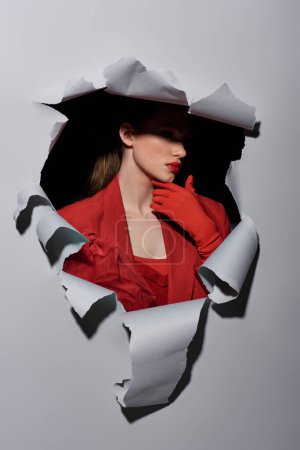 glamorous young woman with bold makeup posing in red gloves near hole in torn grey background