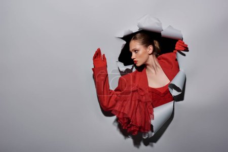 mujer joven con estilo en chaqueta roja y guantes rompiendo a través del agujero en el fondo gris, conceptual