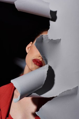 vista parcial de mujer joven con labios rojos posando cerca de papel gris roto, maquillaje audaz y moda