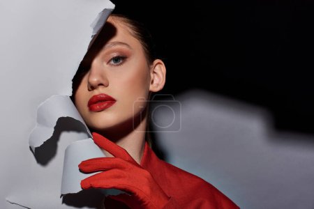 attraktive junge Frau mit roten Lippen, die neben zerrissenem grauen Papier posiert und in die Kamera schaut, fettes Make-up