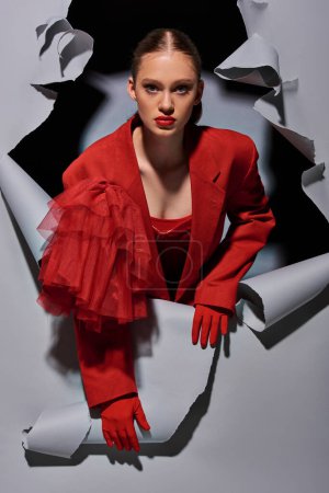 trendy young woman in red attire with bold makeup breaking through grey background with hole