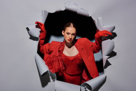 Foto de Poderosa mujer joven en traje rojo con maquillaje audaz rompiendo a través de fondo gris con agujero - Imagen libre de derechos