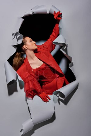 stylish young woman in red attire with bold makeup breaking through grey background with hole