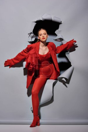 fashionable young woman in red outfit with bold makeup breaking through grey background with hole
