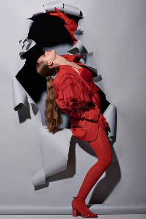 fashionable young woman in red outfit with bold makeup posing near torn grey background with hole