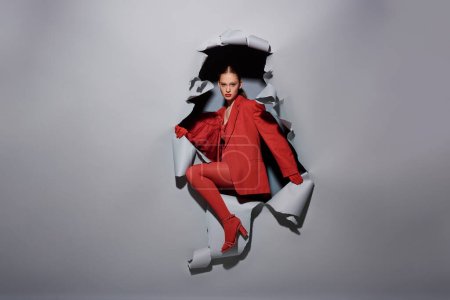 powerful young woman in red outfit with bold makeup breaking through torn grey background with hole