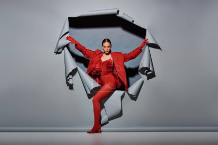 confident young woman in red outfit with gloves breaking through torn grey background with hole