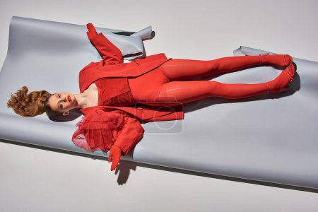 top view of pretty young model in red outfit lying on grey background, woman acting like a doll