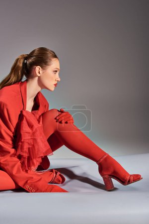 Foto de Vista lateral de la elegante modelo joven en traje rojo con medias y tacones altos sentados sobre fondo gris - Imagen libre de derechos