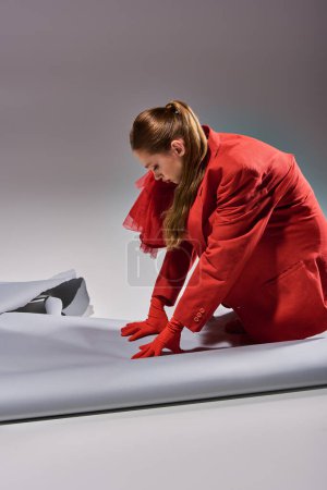 jeune femme en tenue rouge avec gants et talons hauts assis près du papier déchiré sur fond gris