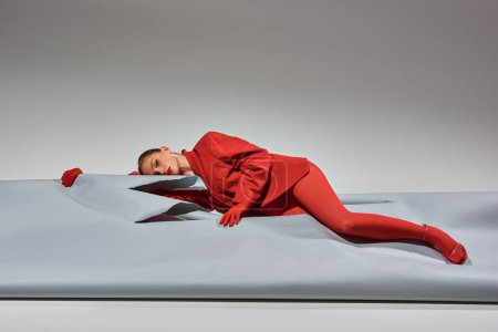 young woman in red attire with gloves and tights lying near ripped paper on grey background